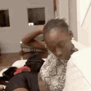 A Man Is Sitting On A Bed In A Living Room With His Hands On His Head . GIF