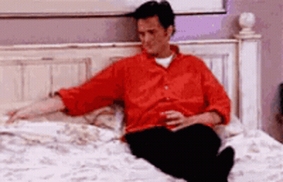 A Man Is Sitting On A Bed With A Drink In His Hand . GIF