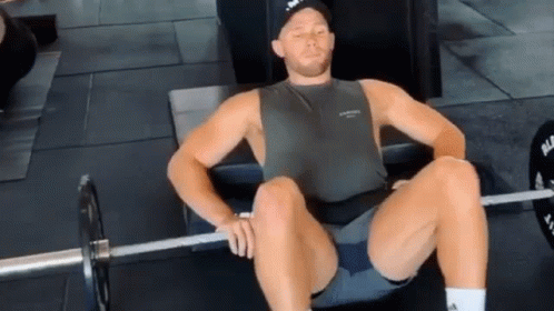 A Man Is Sitting On A Bench With A Barbell In His Lap . GIF