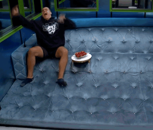 A Man Is Sitting On A Blue Couch With A Bowl Of Strawberries On A Small Table GIF