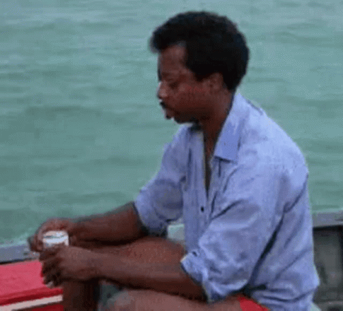 A Man Is Sitting On A Boat In The Water Holding A Can Of Beer . GIF