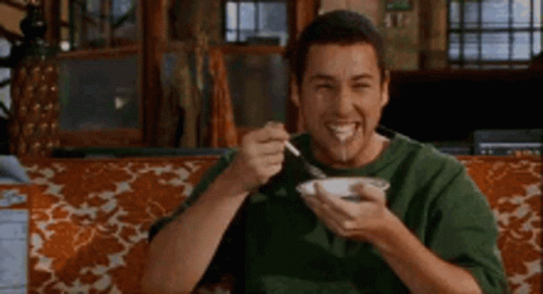 A Man Is Sitting On A Couch Eating A Bowl Of Food With A Fork . GIF