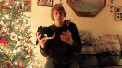 A Man Is Sitting On A Couch Holding A Stuffed Animal In Front Of A Christmas Tree GIF