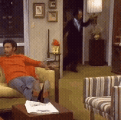 A Man Is Sitting On A Couch In A Living Room While A Man In A Suit Is Standing In The Doorway . GIF