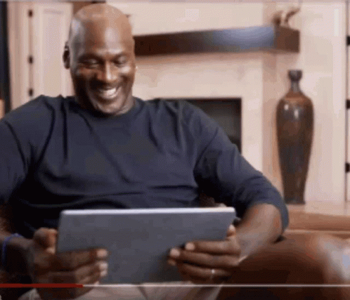 A Man Is Sitting On A Couch Looking At A Tablet And Smiling . GIF