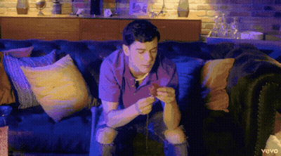 A Man Is Sitting On A Couch Looking At His Phone With The Word Vevo On The Bottom Left GIF