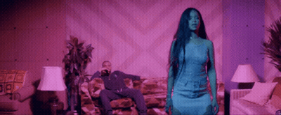 A Man Is Sitting On A Couch While A Woman In A Blue Dress Stands In Front Of Him . GIF
