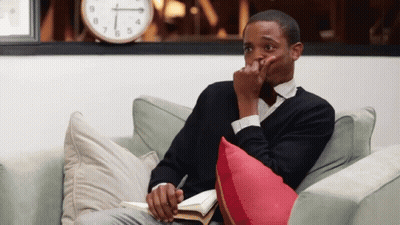 A Man Is Sitting On A Couch With A Book And A Clock Behind Him That Says 1:15 GIF