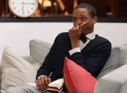 A Man Is Sitting On A Couch With A Book And A Clock Behind Him GIF