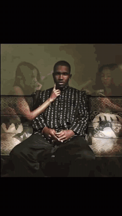 A Man Is Sitting On A Couch With A Woman Behind Him Holding His Hand To His Mouth GIF
