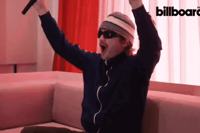 A Man Is Sitting On A Couch With His Arms In The Air In Front Of A Billboard Ad GIF