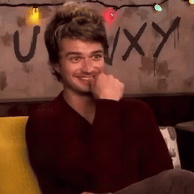 A Man Is Sitting On A Couch With His Hand To His Mouth And A Sign In The Background That Says Uxy . GIF
