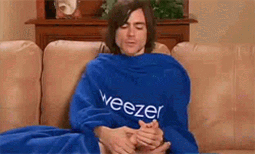 A Man Is Sitting On A Couch Wrapped In A Blue Blanket With The Word Weezer On It . GIF