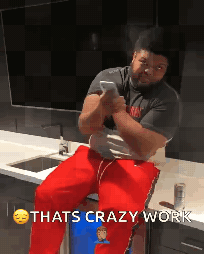 A Man Is Sitting On A Counter Looking At His Phone With The Words Thats Crazy Work Above Him GIF