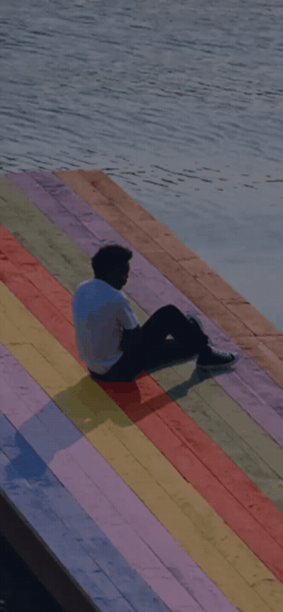 A Man Is Sitting On A Rainbow Colored Dock Overlooking A Body Of Water GIF