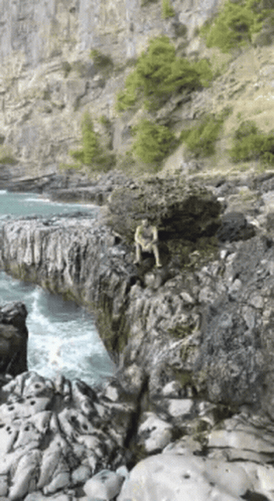 A Man Is Sitting On A Rock Near The Water . GIF