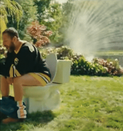 A Man Is Sitting On A Toilet In The Grass With A Sprinkler Spraying Water GIF
