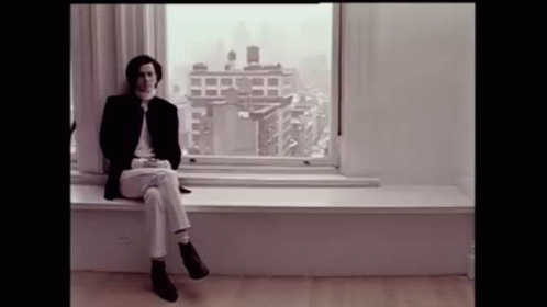 A Man Is Sitting On A Window Sill In Front Of A Window With A View Of A City . GIF