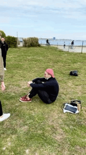 A Man Is Sitting On The Grass In A Park Next To A Body Of Water . GIF