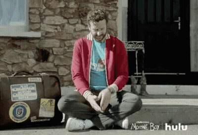 A Man Is Sitting On The Ground In Front Of A Stone Building . GIF