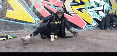 A Man Is Sitting On The Ground In Front Of A Wall With Graffiti On It That Says ' Fun ' GIF