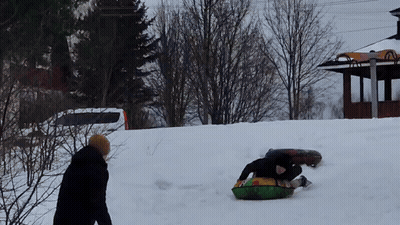A Man Is Sledding Down A Snow Covered Hill While Another Man Watches GIF