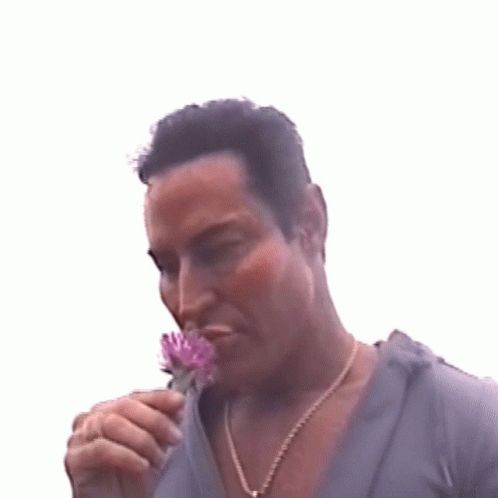 A Man Is Smelling A Purple Flower While Wearing A Gold Chain Around His Neck GIF