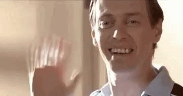A Man Is Smiling And Waving His Hand At The Camera . GIF