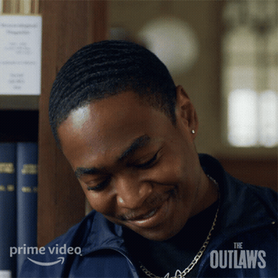 A Man Is Smiling In Front Of A Bookshelf With The Outlaws Written On The Bottom Right Corner GIF