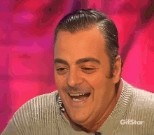 A Man Is Smiling In Front Of A Pink Background With Gifstar On The Bottom GIF