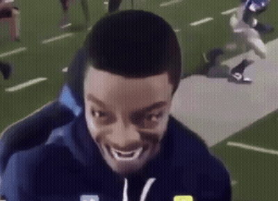 A Man Is Smiling On A Football Field While A Football Player Is Running Behind Him . GIF