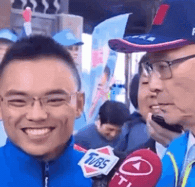 A Man Is Smiling While Talking Into A Microphone While Wearing Glasses And A Hat . GIF