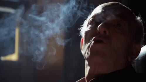 A Man Is Smoking A Cigarette In A Dark Room With Smoke Coming Out Of His Mouth GIF