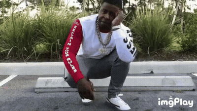 A Man Is Squatting Down In A Parking Lot While Wearing A White Shirt With Arabic Writing On The Sleeves . GIF