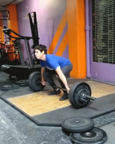 A Man Is Squatting Down To Lift A Barbell In A Gym With A Purple Wall Behind Him GIF