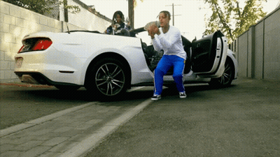 A Man Is Squatting In Front Of A White Car Holding A Bag Of Money GIF