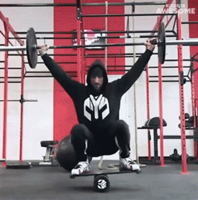 A Man Is Squatting On A Balance Board While Holding A Barbell Over His Head In A Gym GIF
