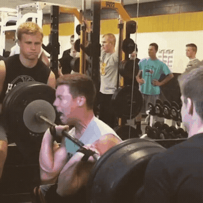 A Man Is Squatting With A Barbell In A Gym While Another Man Looks On GIF