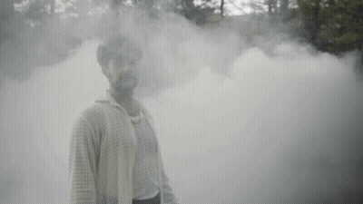 A Man Is Standing In A Foggy Forest And Smiling At The Camera . GIF
