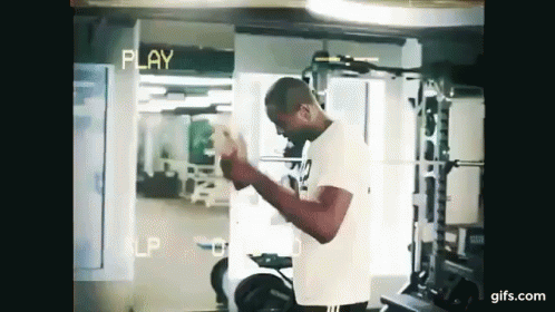 A Man Is Standing In A Gym Holding A Dumbbell In His Hands . GIF