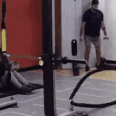 A Man Is Standing In A Gym Next To A Machine With A Rope Attached To It . GIF