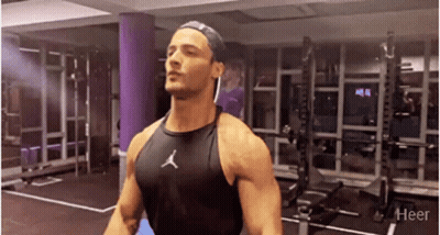 A Man Is Standing In A Gym Wearing A Black Tank Top And A Hat . GIF
