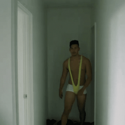 A Man Is Standing In A Hallway Wearing A Yellow Slingshot Underwear . GIF