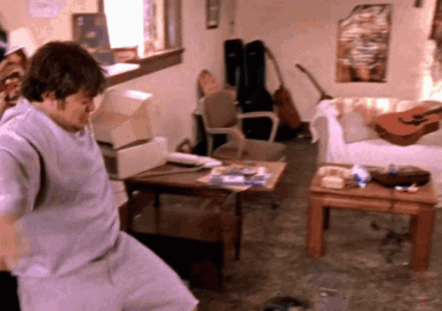 A Man Is Standing In A Living Room With A Guitar On A Table GIF