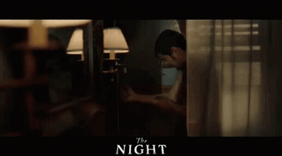 A Man Is Standing In A Room Behind A Curtain And Looking Out A Window . GIF