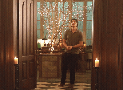 A Man Is Standing In A Room With Candles And A Tree With Lights On It GIF