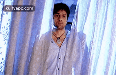 A Man Is Standing In Front Of A Blue Curtain And Looking At The Camera . GIF