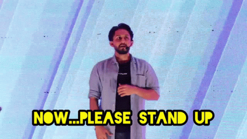 A Man Is Standing In Front Of A Blue Wall And Says Now Please Stand Up GIF
