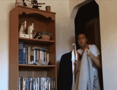 A Man Is Standing In Front Of A Bookshelf With A Book Titled ' A Dictionary ' On The Top Shelf GIF