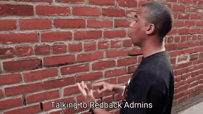 A Man Is Standing In Front Of A Brick Wall Talking To Redback Admins GIF